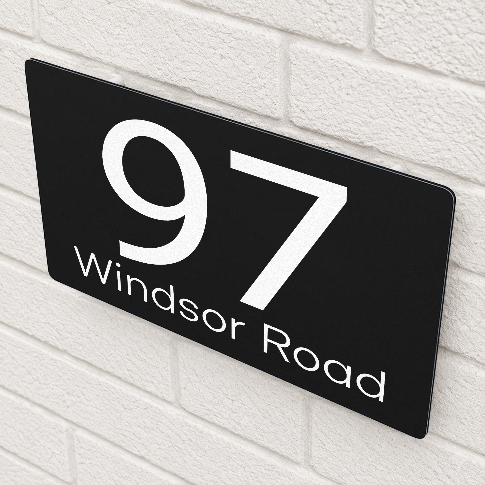 Personalised black house sign reading '97 Windsor Road' viewed at an angle on a textured white brick wall