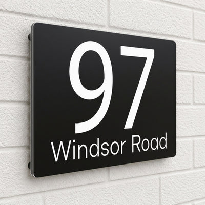 Black personalised house number sign reading '97 Windsor Road' mounted on a white brick wall - SignStation composite aluminium house sign